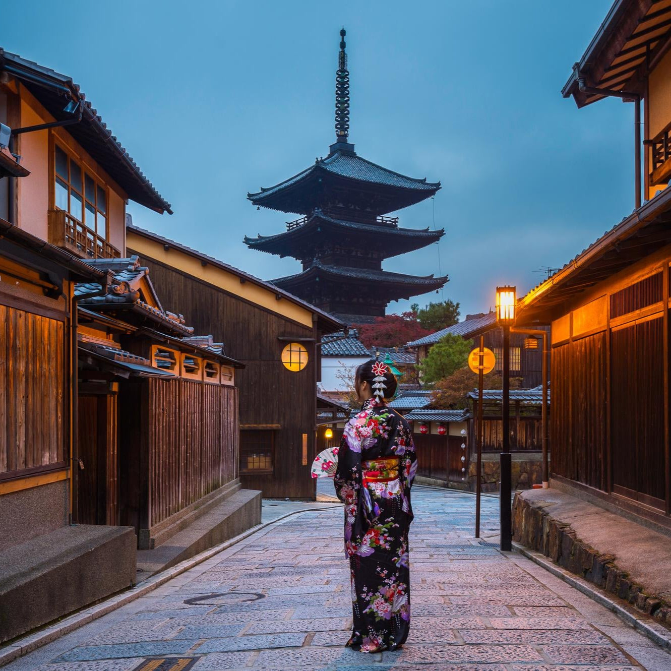 Woman in a traditional kimono standing in a Japanese street with wooden buildings and a pagoda in the background.